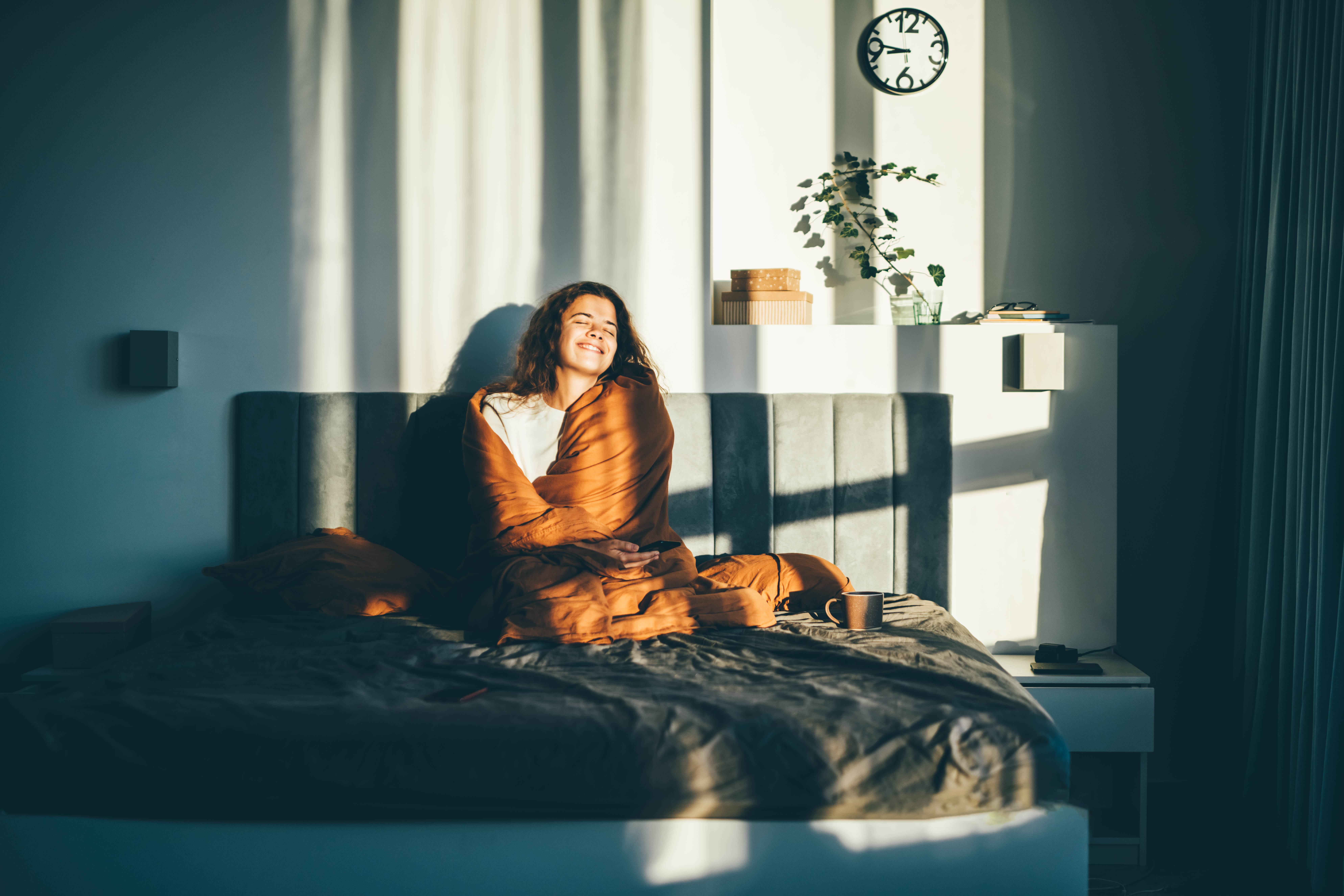 Frau sitzt zugedeckt auf dem Bett und wird von der Sonne angestrahlt