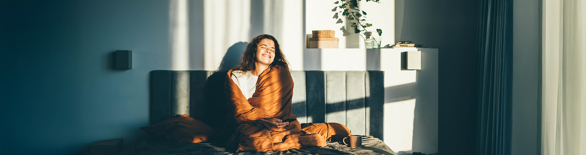 Frau sitzt zugedeckt auf dem Bett und wird von der Sonne angestrahlt